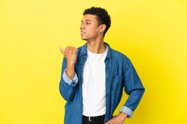 Sarı arka planda izole edilmiş genç Afrikalı Amerikalı bir ürün sunmak için kenarı işaret ediyor.