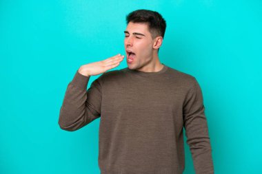 Kafkasyalı genç adam mavi arka planda izole edilmiş esniyor ve geniş açık ağzını eliyle kapatıyor.