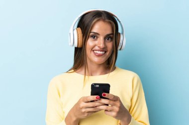 Genç Uruguaylı kadın mavi arka planda izole edilmiş. Cep telefonuyla müzik dinliyor ve ön tarafa bakıyor.