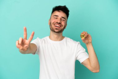 Mavi arka planda izole edilmiş bir Bitcoin tutan genç adam gülümsüyor ve zafer işareti gösteriyor.
