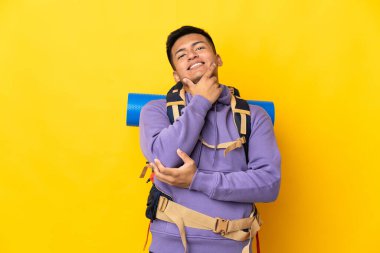 Young mountaineer man with a big backpack isolated on yellow background happy and smiling