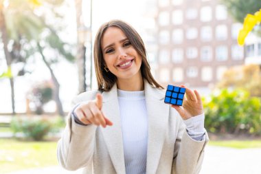 Açık havada üç boyutlu bulmaca küpünü tutan genç ve güzel bir kadın kendinden emin bir ifadeyle seni işaret ediyor.