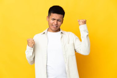 Sarı arka planda izole edilmiş genç Ekvadorlu bir adam zaferi kutluyor.