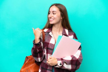 Mavi arka planda izole edilmiş genç beyaz kadın bir ürünü sunmak için kenarı işaret ediyor.