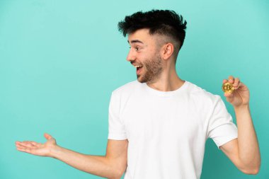 Mavi arka planda Bitcoin tutan genç bir adam sürpriz bir ifadeyle yan yana bakarken