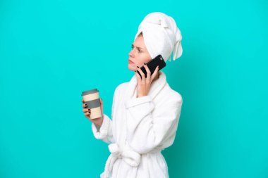 Bornozlu genç bir kadın mavi arka planda izole edilmiş. Elinde kahve ve cep telefonu var.