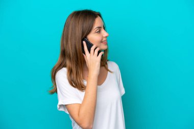 Mavi arka planda izole edilmiş genç beyaz bir kadın cep telefonuyla sohbet ediyor.