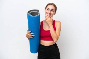 Young sport caucasian woman going to yoga classes while holding a mat isolated on white background looking up while smiling