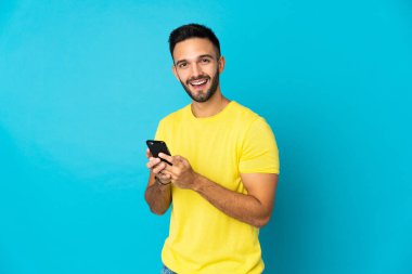 Mavi arka planda izole edilmiş genç beyaz adam cep telefonuyla bir mesaj gönderiyor.