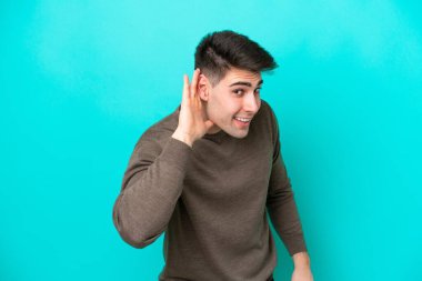 Kafkasyalı genç adam mavi arka planda izole edilmiş bir şekilde elini kulağına koyarak bir şeyler dinliyor.
