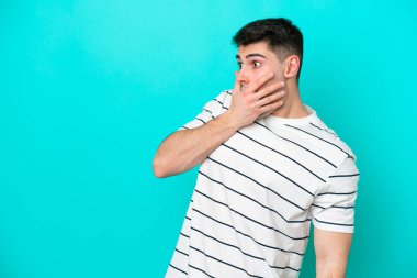 Mavi arka planda izole edilmiş genç beyaz adam yan tarafa bakarken sürpriz jest yapıyor.