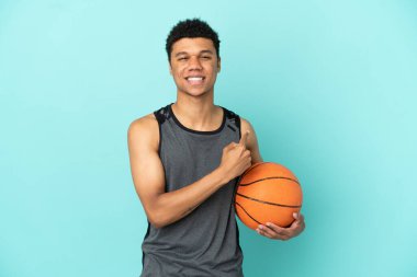 Basketbolcu Afrikalı Amerikalı bir adam mavi arka planda izole edilmiş bir şekilde bir ürünü sunmak için kenarı işaret ediyor.