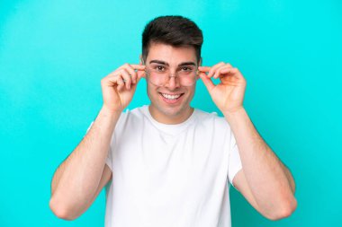 Genç beyaz adam mavi arka planda izole edilmiş, mutlu bir ifadeyle gözlüklü.