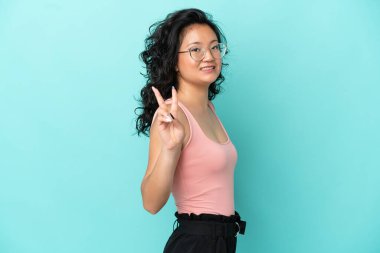 Mavi arka planda izole edilmiş genç Asyalı kadın gülümsüyor ve zafer işareti gösteriyor.