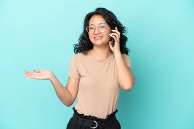 Genç Asyalı bir kadın mavi arka planda izole edilmiş. Cep telefonuyla biriyle sohbet ediyor.