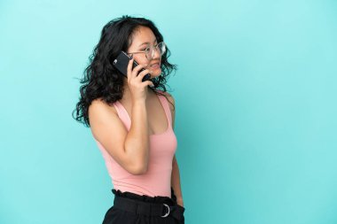 Genç Asyalı bir kadın mavi arka planda izole edilmiş. Cep telefonuyla biriyle sohbet ediyor.