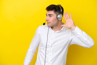 Tele pazarlamacı beyaz adam sarı arka planda izole edilmiş bir kulaklıkla çalışıyor. Kulağına elini sürerek bir şeyler dinliyor.