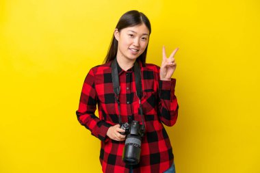 Sarı arka planda izole edilmiş genç fotoğrafçı kadın gülümsüyor ve zafer işareti gösteriyor.