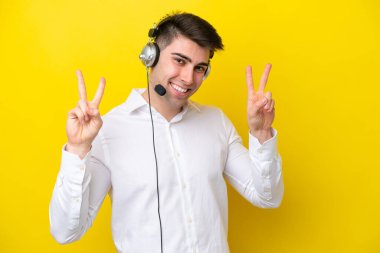 Tele pazarlamacı beyaz adam sarı arka planda izole edilmiş bir kulaklıkla çalışıyor. Zafer işaretini iki eliyle gösteriyor.