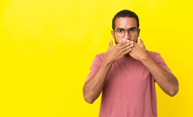 Sarı arka planda izole edilmiş genç latin adam elleriyle ağzını kapatıyor.