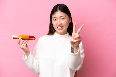 Pembe arka planda saşimi tutan genç Çinli kadın gülümsüyor ve zafer işareti gösteriyor.