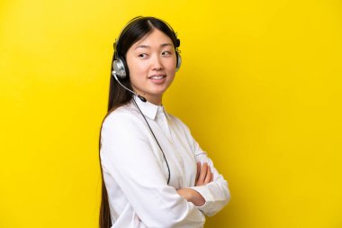 Sarı arka planda kolları bağlı ve mutlu bir şekilde kulaklıkla çalışan pazarlamacı Çinli kadın.