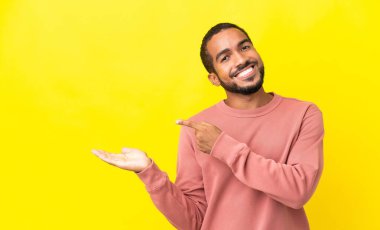 Sarı arka planda izole edilmiş genç latin adam bir ilan vermek için avucunun içinde hayali bir alan tutuyor.