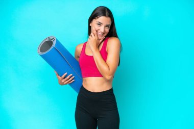Genç beyaz kadın yoga derslerine giderken mavi arka planda izole edilmiş bir minderi tutarken mutlu ve gülümsüyor.