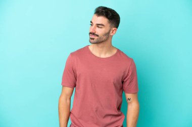 Kafkasyalı genç adam mavi arka planda izole edilmiş, yana bakıyor ve gülümsüyor.