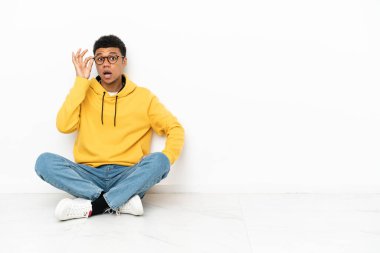 Genç Afrikalı Amerikalı bir adam beyaz arka planda gözlüklü ve şaşırmış bir şekilde yerde oturuyor.