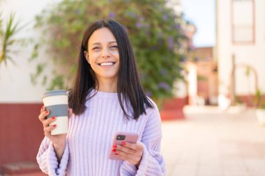 Dışarıda cep telefonu kullanan ve elinde mutlu bir ifadeyle kahve tutan genç bir kadın.