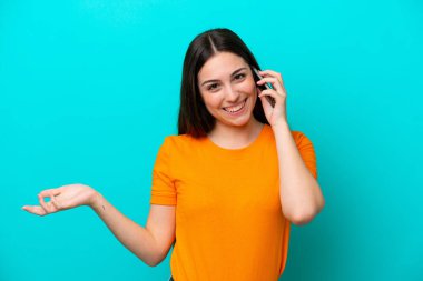 Mavi arka planda izole edilmiş genç beyaz bir kadın cep telefonuyla sohbet ediyor.