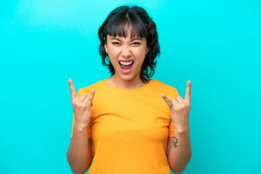 Arjantinli genç bir kadın mavi arka planda izole edilmiş boynuz hareketi yapıyor.