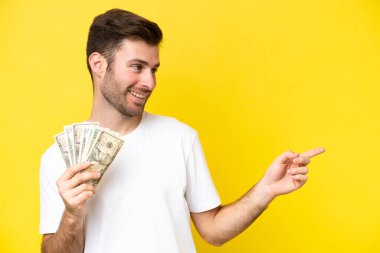 Kafkasyalı genç adam sarı arka planda izole edilmiş bir sürü para alıyor. Parmakla işaret ediyor ve bir ürün sunuyor.