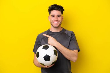 Yakışıklı genç futbolcu sarı arka planda izole edilmiş bir şekilde bir ürün sunmak için kenarı işaret ediyor.