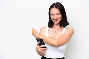 Beyaz arka planda izole edilmiş genç beyaz bir kadın cep telefonu kullanıyor ve geriye doğru işaret ediyor.