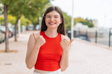 Açık havada gözlüklü genç bir kadın. Baş parmakları havada ve gülümsüyor.