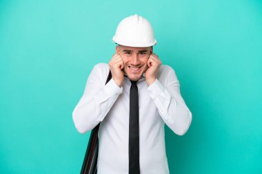 Kasklı ve elinde mavi arka planda izole edilmiş planları olan genç bir beyaz adam hayal kırıklığına uğramış ve kulaklarını kapatmış.