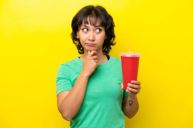 Young Argentinian woman holding a soda isolated on yellow background having doubts