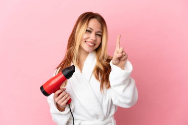 İzole edilmiş pembe arka planda saç kurutma makinesi tutan genç sarışın kız en iyinin işareti olarak parmağını kaldırıyor.