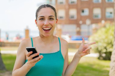 Açık havada cep telefonu kullanan genç ve güzel bir kadın şaşırmış ve parmağıyla kenarı işaret ediyor.