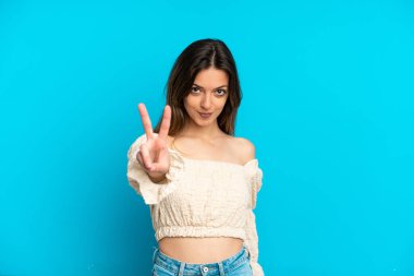 Mavi arka planda izole edilmiş genç beyaz kadın gülümsüyor ve zafer işareti gösteriyor.