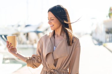 Açık havada elinde kahveyle gezen genç bir kadın. Mutlu bir ifadeyle.