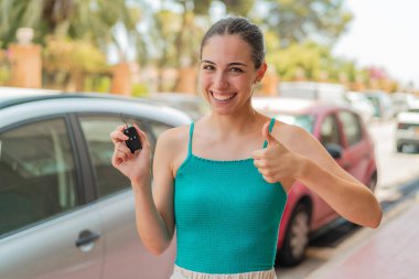 Dışarıda arabanın anahtarlarını tutan genç ve güzel bir kadın. Başparmakları havada çünkü iyi bir şey oldu.