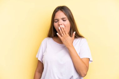 Sarı arka planda izole edilmiş genç, beyaz bir kadın esniyor ve ağzını eliyle kapatıyor.