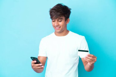 Kafkasyalı genç adam mavi arka planda izole edilmiş cep telefonuyla kredi kartıyla alışveriş yapıyor.