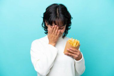 Elinde kızarmış cipsler tutan genç Arjantinli kadın yorgun ve hasta bir ifadeyle mavi arka planda izole edilmiş.