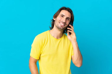 Genç ve yakışıklı bir adam mavi arka planda izole edilmiş, cep telefonuyla sohbet ediyor.
