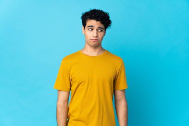 Genç Venezuelalı adam mavi arka planda izole edilmiş. Şüpheli hareketler yapıyor.