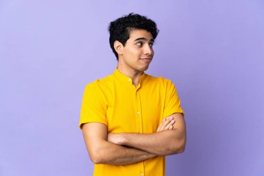 Genç Venezuelalı bir adam mor arka planda izole edilmiş. Omuzlarını kaldırırken şüpheyle hareket ediyor.